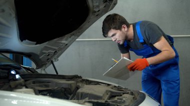 Mechanic examines the motor of car and takes notes