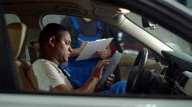 Mechanic with a tablet sits in car and tells to colleague to take notes
