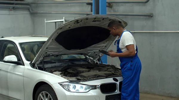 Mechanic in uniform repairs a car and types on tablet — Stockfoto