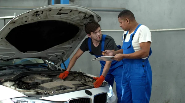 Mechanic in uniform repairs a car, his collegue makes notes — Stockfoto