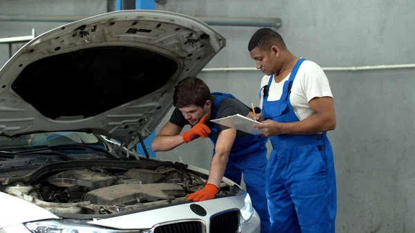 Mechanic in uniform repairs a car, his collegue types on tablet — Stock Photo, Image