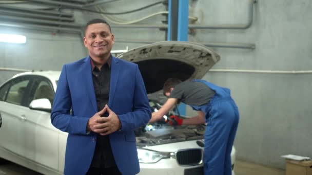 Человек в костюме улыбается в камеру, механик чинит машину на заднем плане — стоковое видео