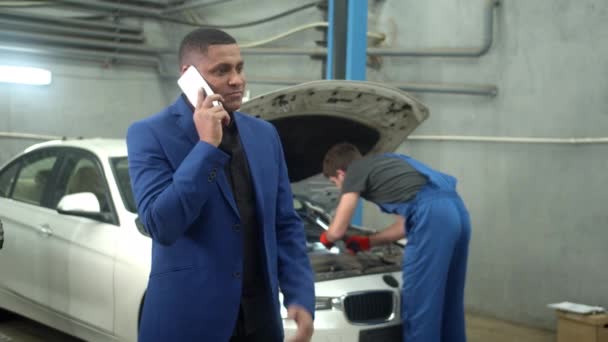 El hombre habla por teléfono, mecánico repara un coche en el fondo, cámara lenta — Vídeo de stock