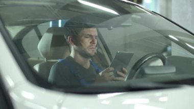 Mechanic sits in a car and looks at something on a tablet