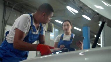 Tamirci araba tamir eder, kadın oto tamirhanesinde elinde tabletle durur.