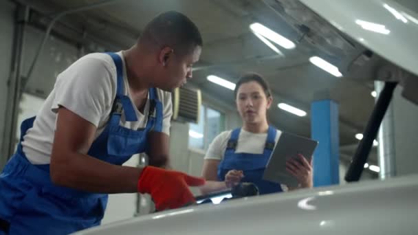 Mecánico repara un coche, mujer se para con la tableta en el taller de reparación de automóviles — Vídeos de Stock