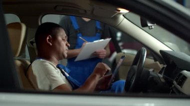 Mechanic with a tablet sits in car and tells to colleague to take notes