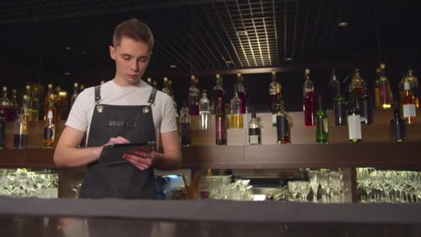 Bartender stands with tablet and counts bottles with alcohol — ストック動画