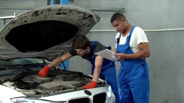 Mechaniker in Uniform repariert Auto, sein Kollege tippt auf Tablet — Stockvideo