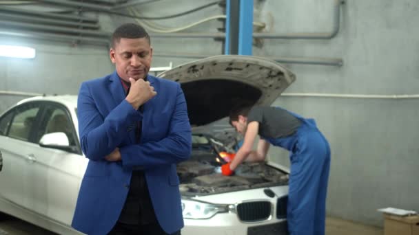 Man i kostym visar missnöje vid kameran, mekaniker reparerar en bil på bakgrunden — Stockvideo