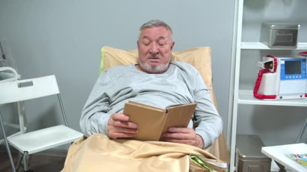 Чоловік лежить на ліжку в лікарні і читає книгу — стокове відео