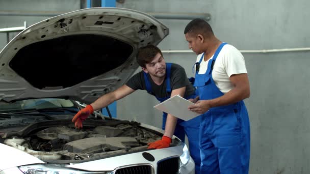 Mechanik v uniformě opravuje auto, jeho kolega si dělá poznámky — Stock video