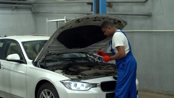 Mecánico en uniforme repara un coche y toma notas — Vídeo de stock