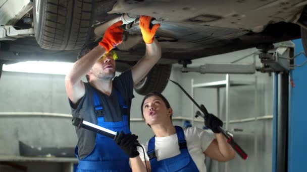 Mechanik w okularach naprawia samochód, kobieta trzyma latarkę — Wideo stockowe