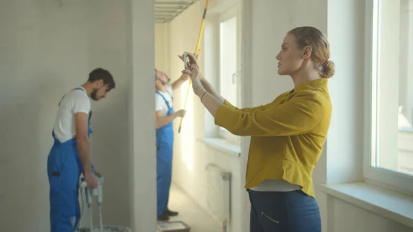 Kadın telefonda fotoğraf çekiyor, tamirciler dairede çalışıyor.