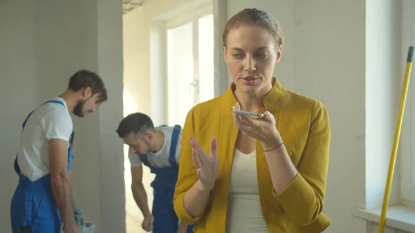 Woman records a voice message on phone, builders in uniform work