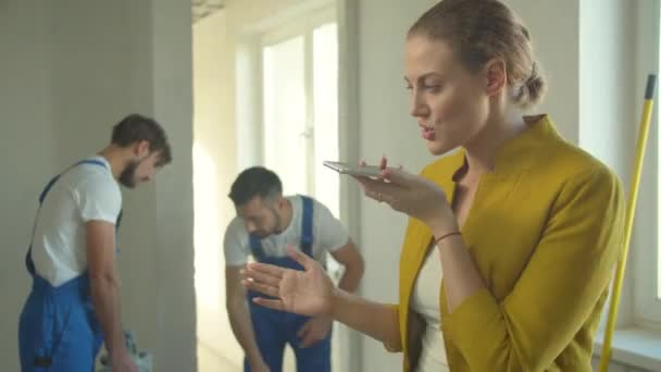 Mulher grava uma mensagem de voz no telefone, construtores em trabalho uniforme — Vídeo de Stock