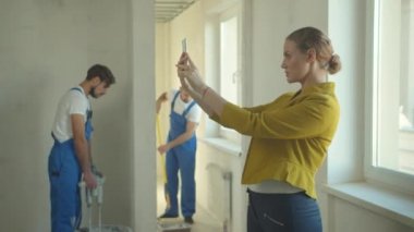 Kadın telefonda fotoğraf çekiyor, tamirciler dairede çalışıyor.