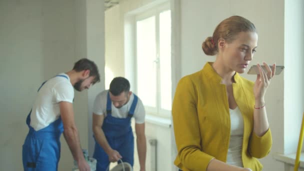 Kvinna spelar in ett röstmeddelande på telefon, byggare arbetar i lägenheten — Stockvideo