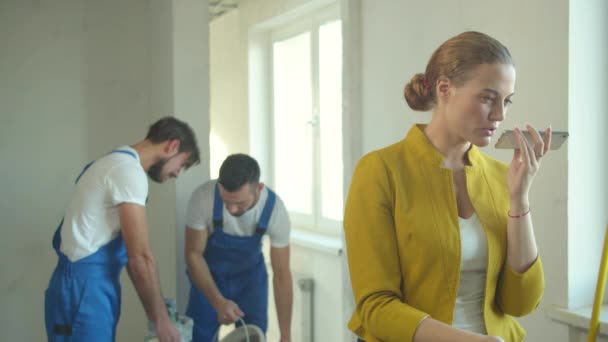 La mujer graba un mensaje de voz en el teléfono, los constructores trabajan en el piso — Vídeo de stock