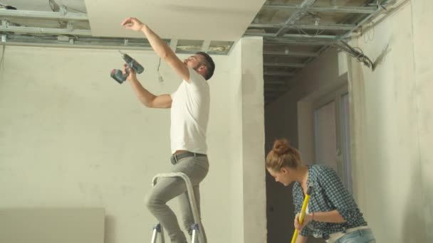 El hombre sujeta el panel en el techo, la mujer pinta la pared — Vídeos de Stock
