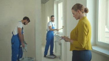 Kadın tabletle duruyor ve inşaatçıların nasıl çalıştığını izliyor. 
