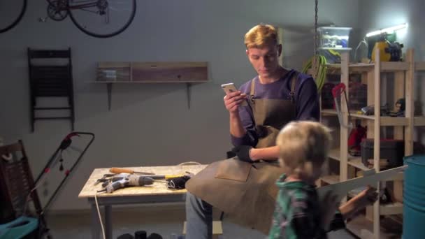 Мужчина сидит с телефоном, мальчик играет с деревянным пистолетом, замедленная съемка — стоковое видео