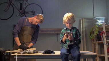 Çocuk telefonuyla oturur, adam atölyede çalışır.