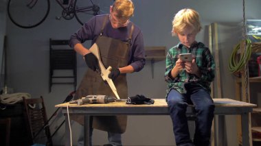 Çocuk telefonuyla oturur, usta atölyede ahşapla çalışır.