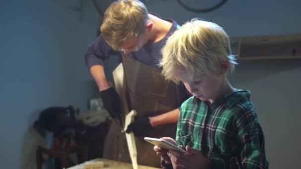 Сын играет в игры по телефону и не мешает отцу работать в гараже — стоковое видео