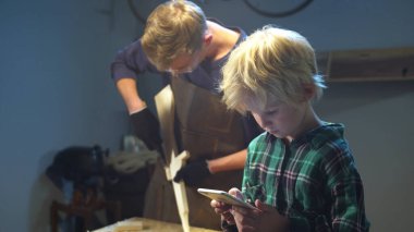 Oğlu telefonda oyun oynuyor ve babasının garajda çalışmasına engel olmuyor.