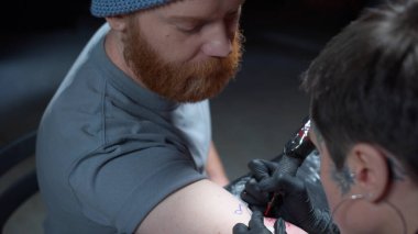 Sakallı bir adam dövmeyi dolduran bir dövmeciyi izliyor.