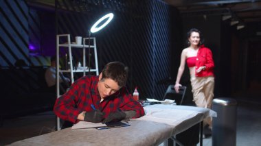 Dövme ustası bir eskiz çiziyor. Model arka planda duruyor.