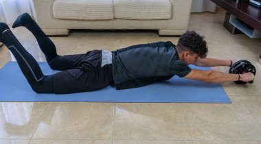 Siyah spor kıyafetli genç adam mavi paspasın üzerinde oturma odasında egzersiz yapıyor.