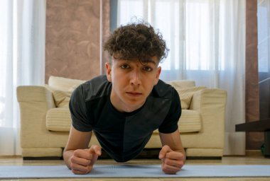 Siyah spor kıyafetli genç adam mavi paspasın üzerinde oturma odasında egzersiz yapıyor.