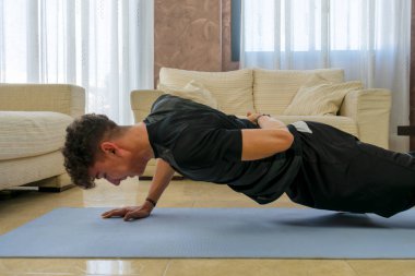 Siyah spor kıyafetli genç adam mavi paspasın üzerinde oturma odasında egzersiz yapıyor.