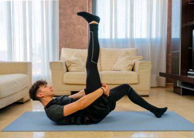 Siyah spor kıyafetli genç adam mavi paspasın üzerinde oturma odasında egzersiz yapıyor.