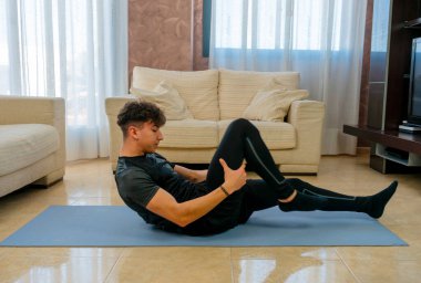 Siyah spor kıyafetli genç adam mavi paspasın üzerinde oturma odasında egzersiz yapıyor.