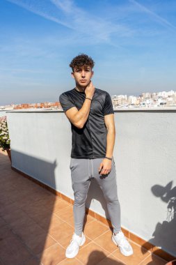 Mavi gökyüzünün altındaki terasta siyah spor kıyafetli genç adam poz veriyor.