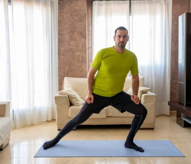 Sakallı adam, siyah ve yeşil spor kıyafetleriyle egzersiz yapıyor oturma odasında minderdeki kanepelerin önünde.