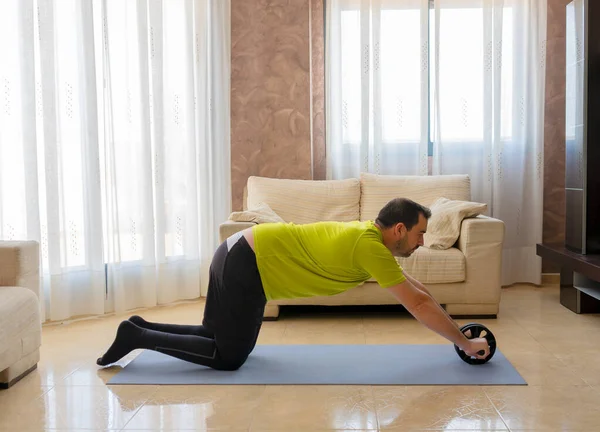 Bearded man in low shape exercising with black and green sportswear in his living room in front of the sofas on a mat