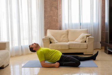 Sakallı adam, siyah ve yeşil spor kıyafetleriyle egzersiz yapıyor oturma odasında minderdeki kanepelerin önünde.