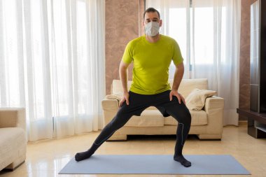 Sakallı adam, siyah ve yeşil spor kıyafetleriyle egzersiz yapıyor ve evdeki paspasın üzerindeki oturma odasında koronavirüsü önlemek için maske takıyor.