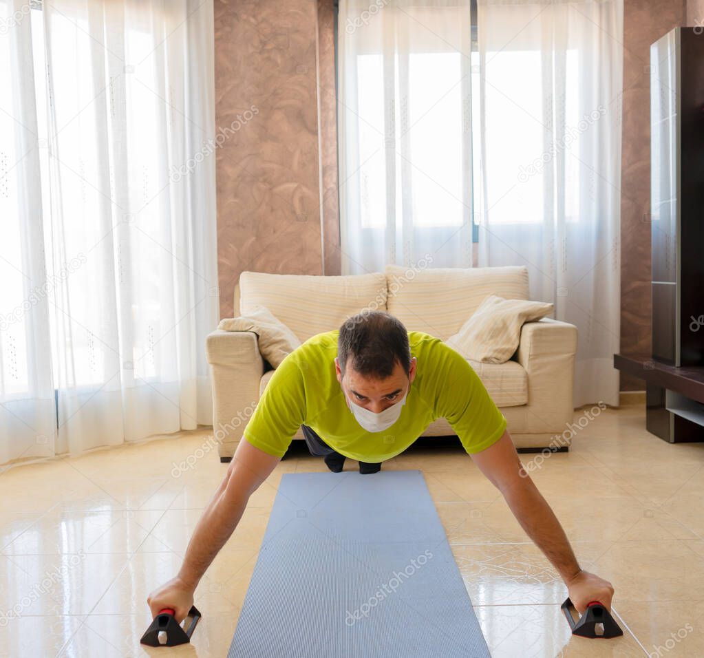 Bearded man in low shape exercising with black and green sportswear and a mask to prevent coronavirus in the living room at home on a mat