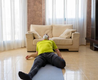 Sakallı adam, siyah ve yeşil spor kıyafetleriyle egzersiz yapıyor ve evdeki paspasın üzerindeki oturma odasında koronavirüsü önlemek için maske takıyor.