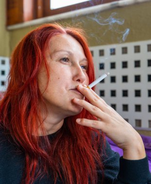 Kızıl saçlı, orta yaşlı güzel bir kadın derin bir tavırla sigara içiyor.