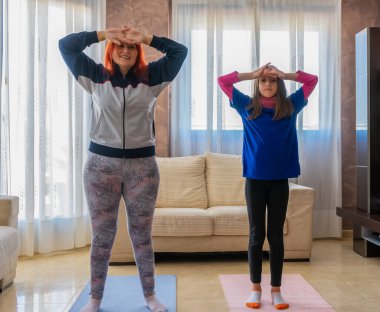 Anne ve kız spor kıyafetleri giyip evdeki oturma odasında beyaz koltukların arasında egzersiz yapıyorlar. Sağlıklı yaşam konsepti