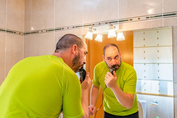 Man with a shaved head and wearing a pistachio green shirt shaving his beard in front of the mirror