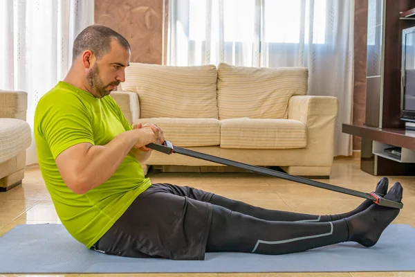 Bearded man in low shape exercising with black and green sportswear in his living room in front of the sofas on a mat