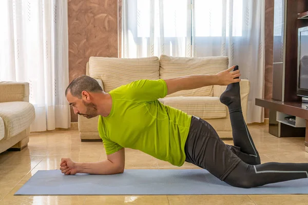 Bearded man in low shape exercising with black and green sportswear in his living room in front of the sofas on a mat